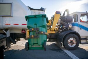 Green wheeled curbside cart being serviced by a side loader truck - suspended in the air by the truck's yellow mechanical arm.