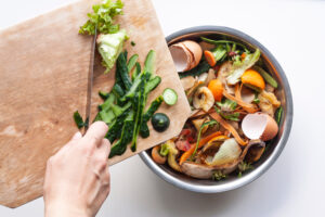A hand scraping various green food scraps into a bowl full of food scraps like eggshells and orange peels