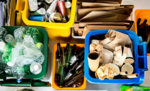 Yellow indoor recycling bin filled with metal cans, Green indoor recycling bin full of plastic bottles, orange indoor recycling bin full of glass bottles, gray indoor recycling bin full of flattened cardboard boxes, and blue indoor recycling bin full of mixed paper.
