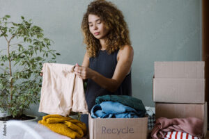 20-30 year old woman with curly hair folding cloth and surrounded with boxes that say "recycle" on them