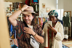 Caucasian young adult man trying on hat and smiling while standing in thrift shop, Black young adult woman standing nearby using smartphone to take photo of him, clothing racks in background